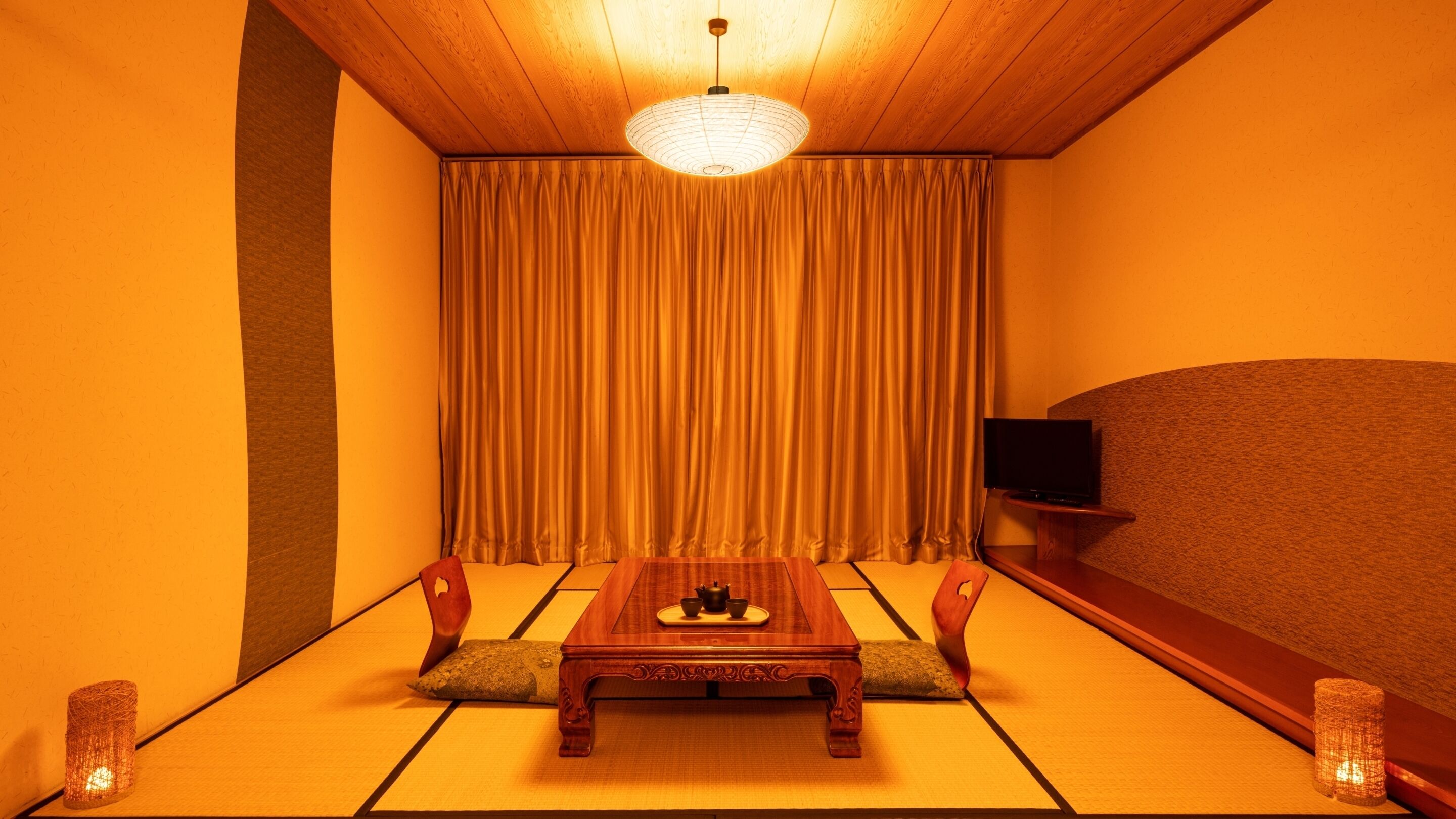 A traditional Japanese room with tatami flooring, a low wooden table with a tea set, a television on a stand, and a sliding door with curtains.