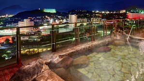 Ett område med varma källor, en pool omgiven av sten, ett glasräcke och utsikt över en stadssiluett i nattljus.