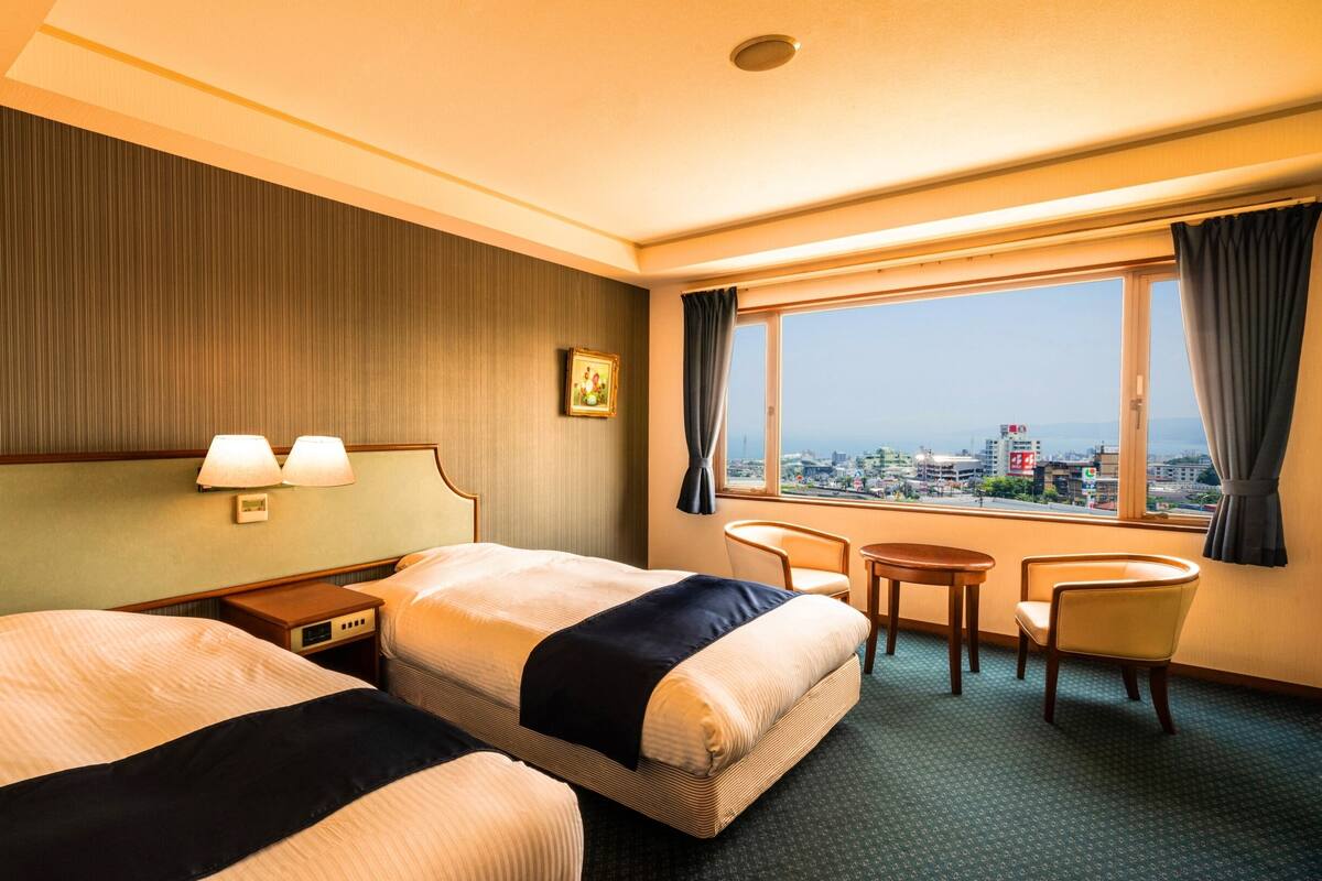 A hotel room with two beds, a small table, a chair, a window with a city view, and a picture on the wall.