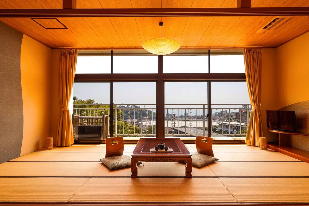 A traditional Japanese room with tatami flooring, a low table with a tea set, and a large window offering a city view.