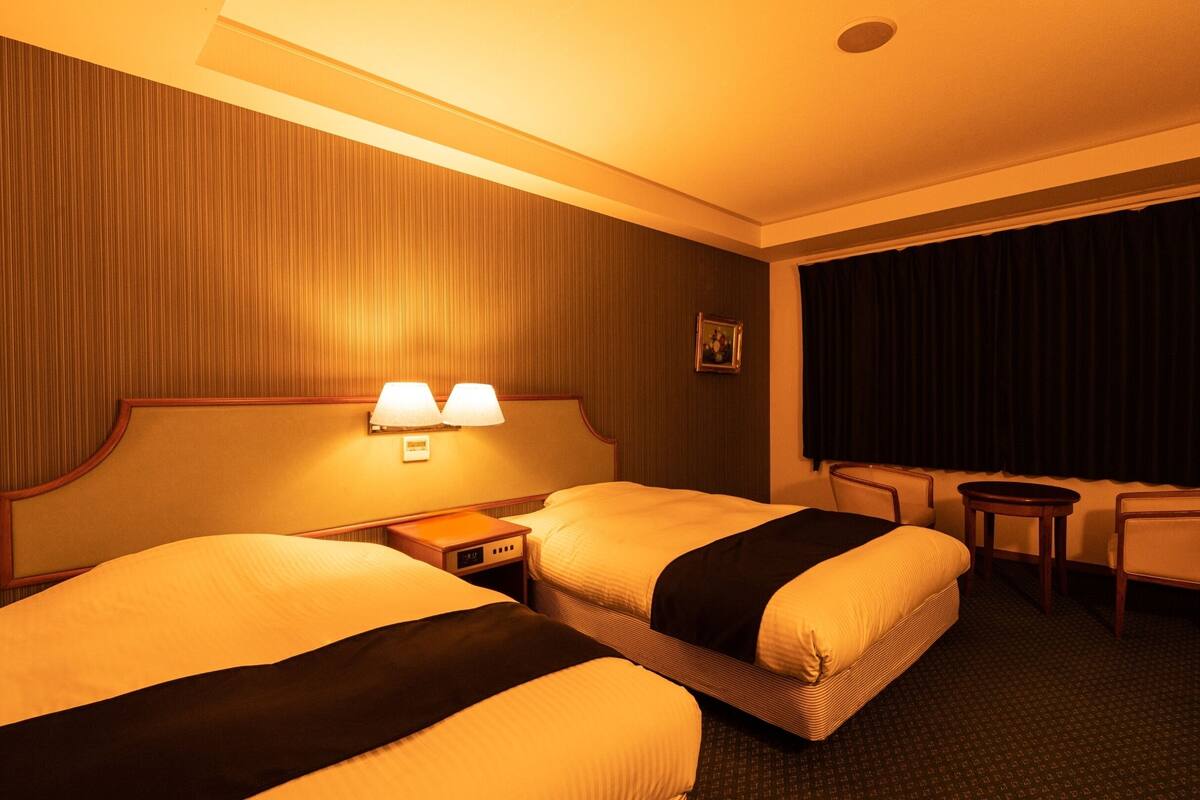 A hotel room with two beds, a nightstand, a lamp, a small table, and a chair.