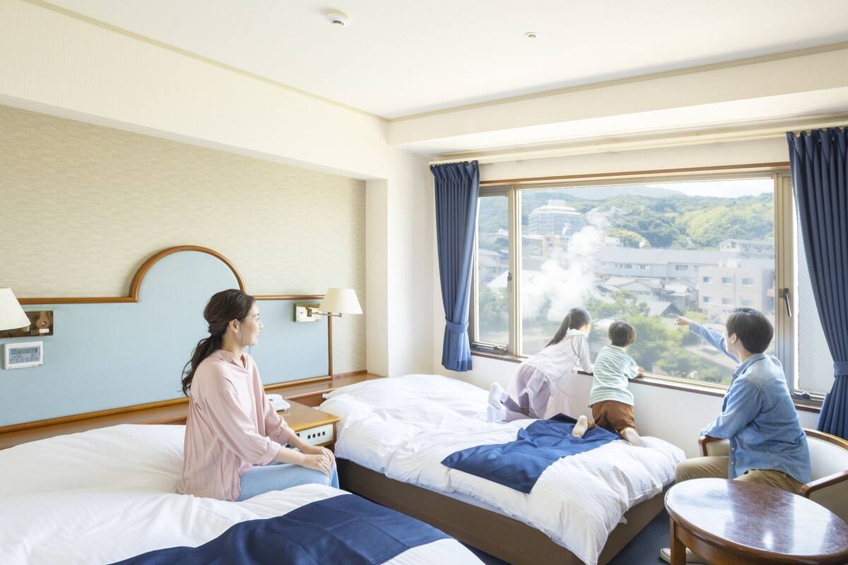 A hotel room with two beds, a small table, and a view of a cityscape.