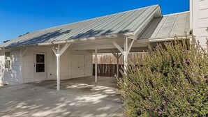 Un espace extérieur abrité, doté d’un toit en métal, un bâtiment blanc avec une porte de garage, et un arbuste aux fleurs violettes.