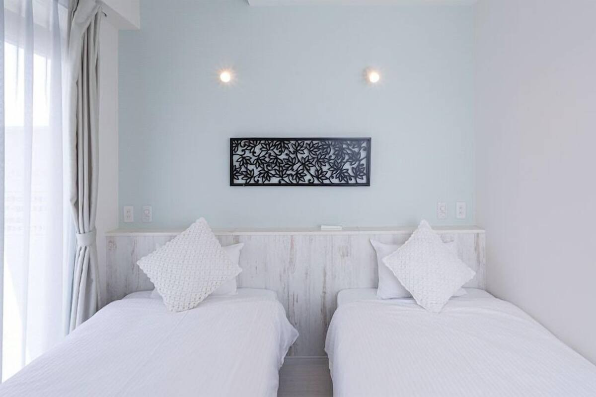 A room with two beds, a light blue wall, a decorative black panel, and a window with curtains.