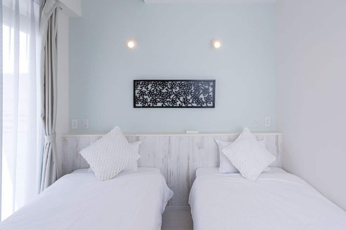 Quarto com duas camas, parede azul clara, painel preto decorativo e janela com cortinas.