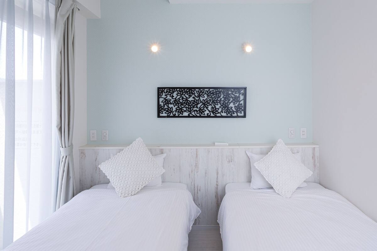 A hotel room with two beds, a light blue wall, a decorative black and white artwork, and a window with sheer curtains.