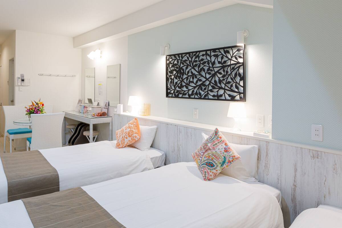 A hotel room with two beds, a desk, a chair, a mirror, and a decorative wall piece.