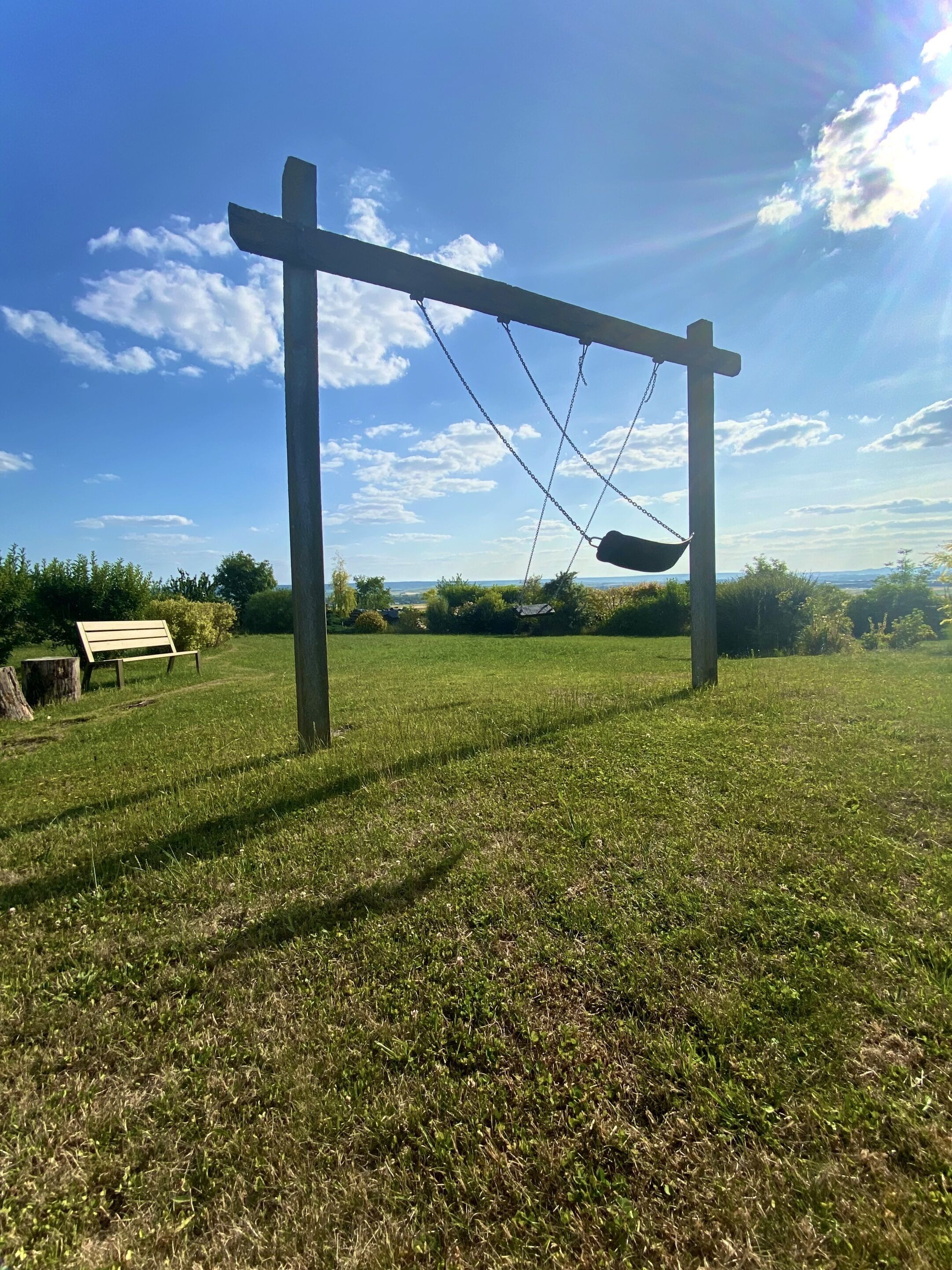 A swing set in a grassy field with a bench and a tree stump nearby.
