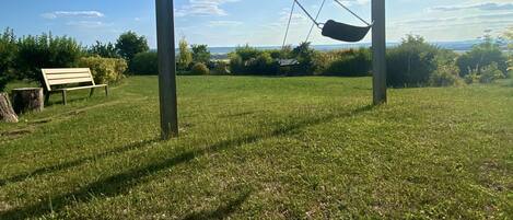 A swing set in a grassy field with a bench and a tree stump nearby.