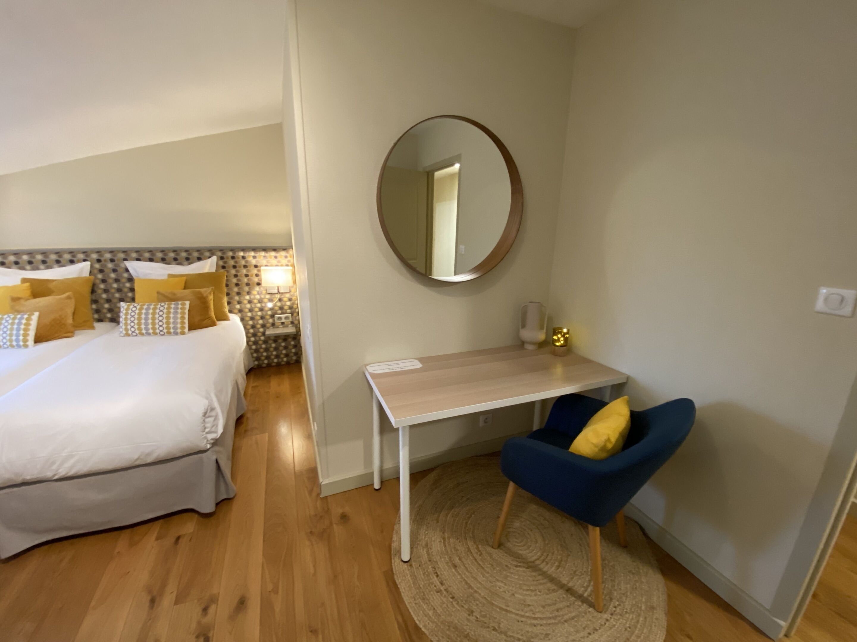 Een hotelkamer met een bed, nachtkastje, stoel en een ronde spiegel aan de muur.