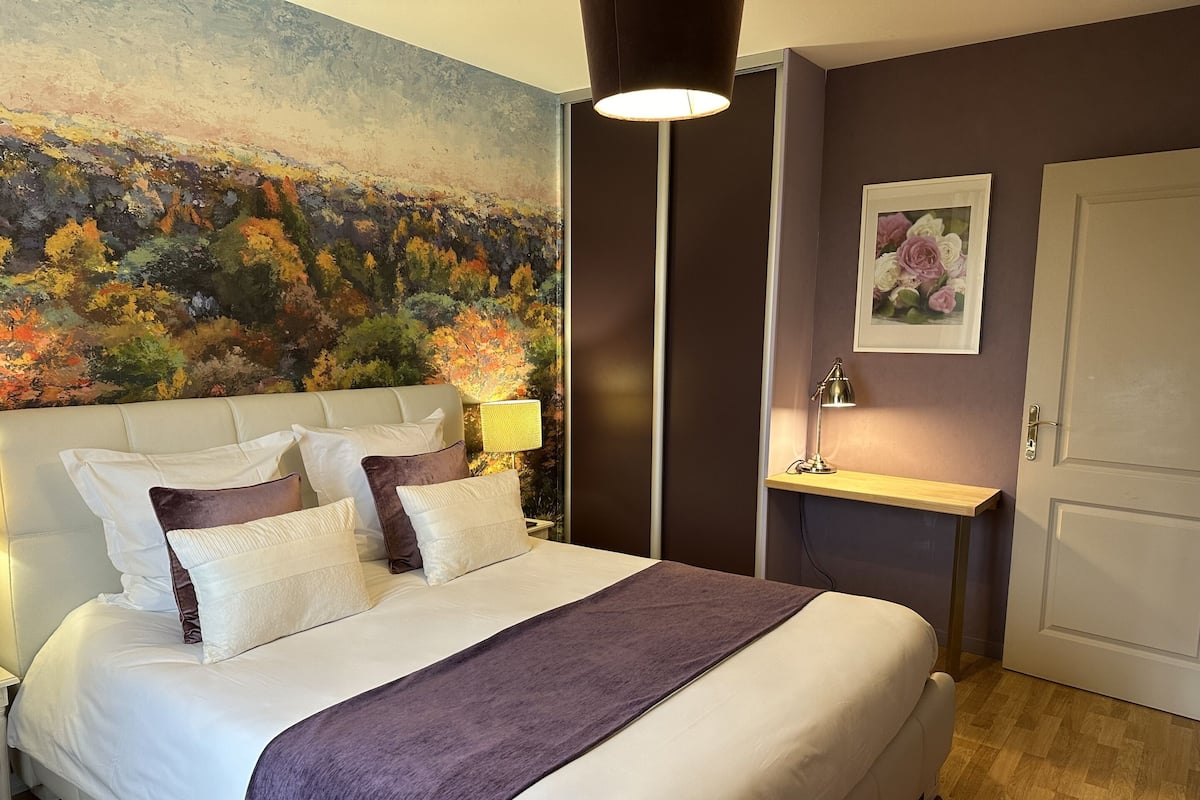 A neatly made bed with a headboard, a bedside table with a lamp, a painting on the wall, and a door.