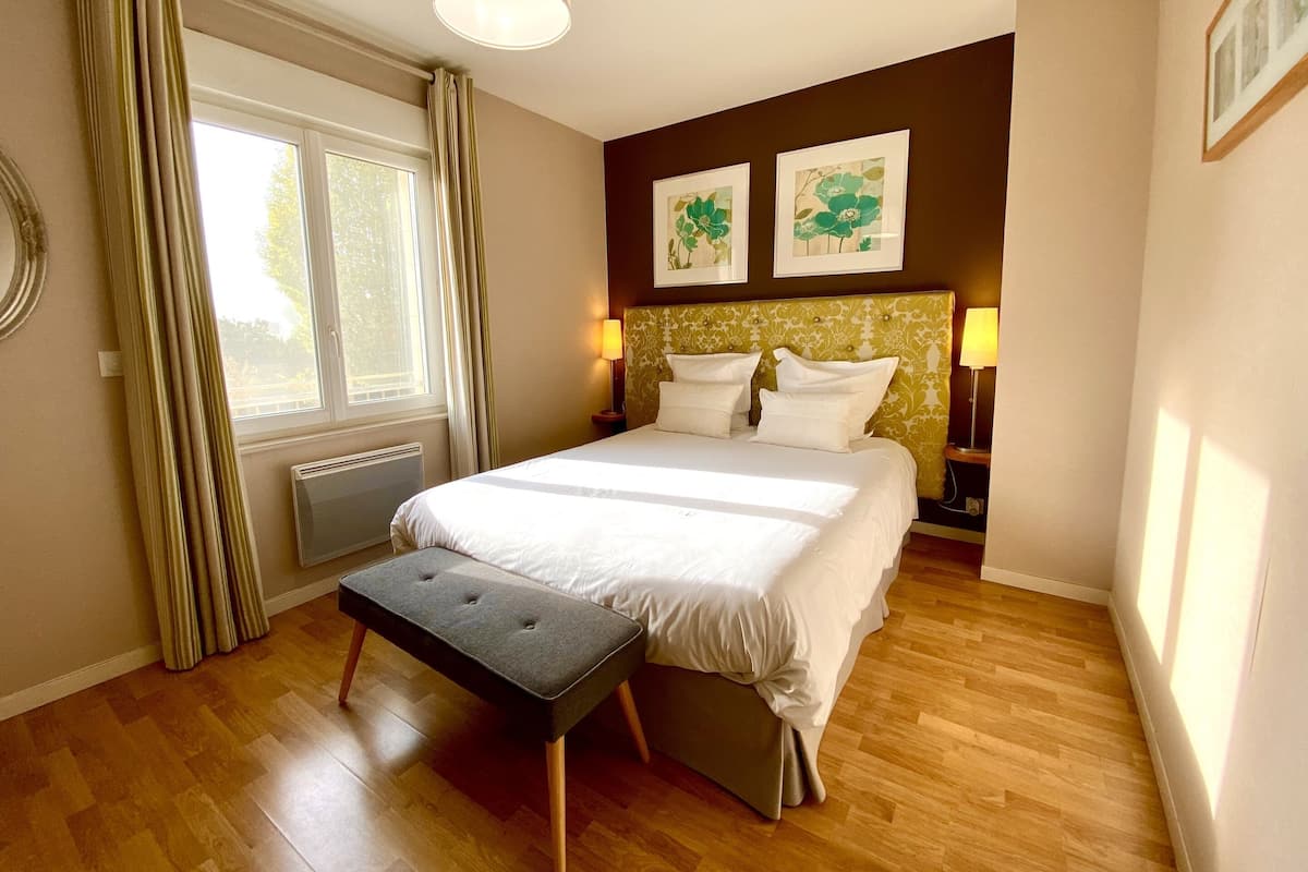 A neatly arranged bedroom with a large bed, a bedside table with lamps, a window with curtains, and two framed floral artworks on the wall.