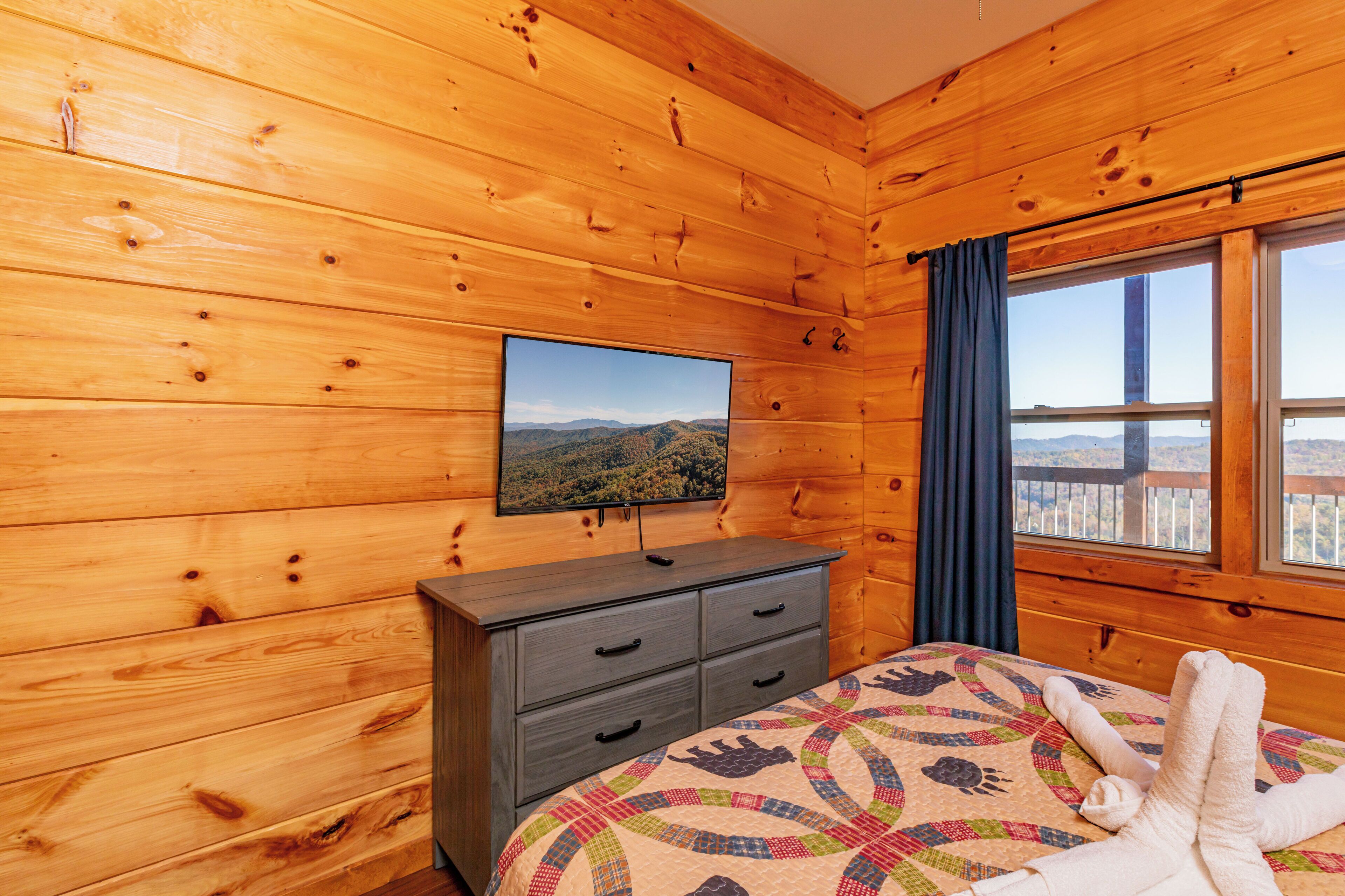 A wooden cabin room with a bed, a dresser, a television on the wall, and a window with a view of the mountains.