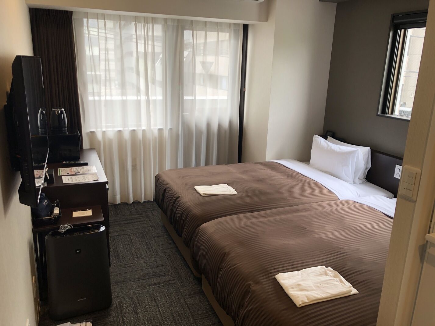 Quarto de hotel com duas camas, uma mesa de trabalho com televisão e uma janela com cortinas.