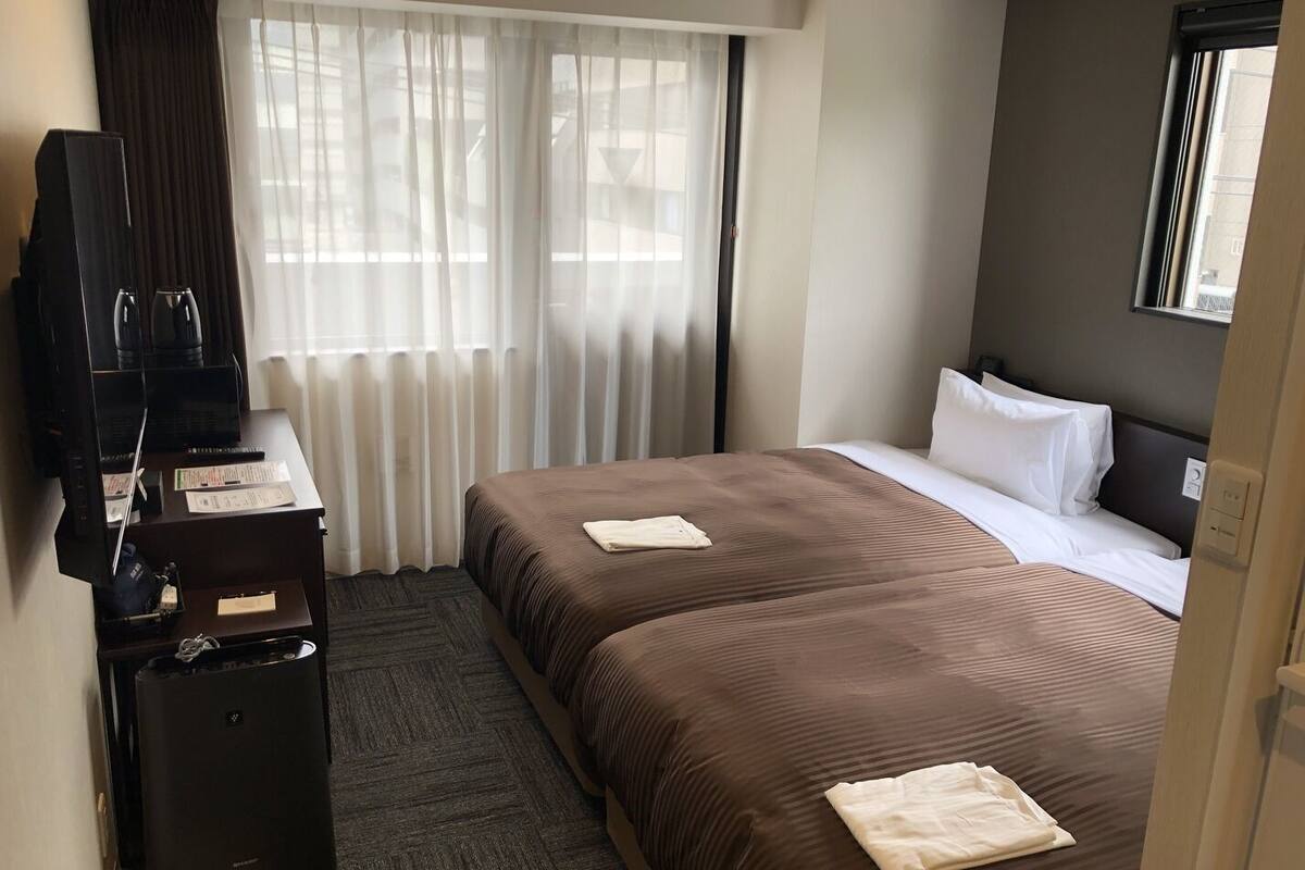 Ein Hotelzimmer mit zwei Betten, einem Schreibtisch mit Fernseher und einem Fenster mit Vorhängen.