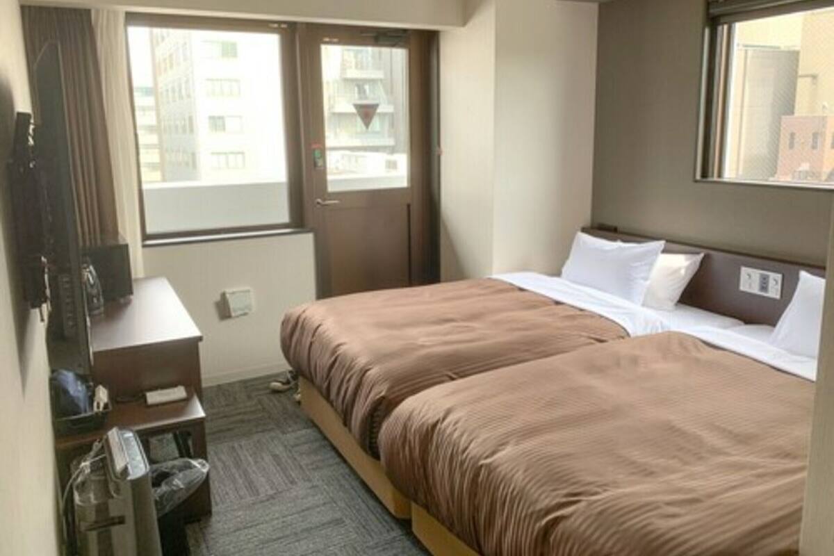 Ein Hotelzimmer mit zwei Betten, einem Schreibtisch, einem Stuhl und einem Fenster mit Blick auf Gebäude.