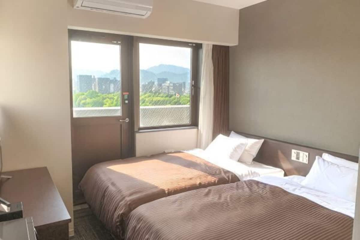 A hotel room with two beds, a desk, and a window with a city view.
