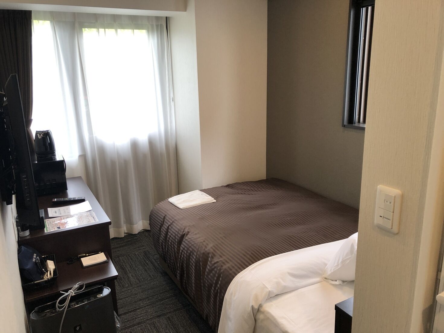 Quarto de hotel com uma cama, uma escrivaninha com um computador e uma cafeteira, uma janela com cortinas e uma televisão fixada na parede.