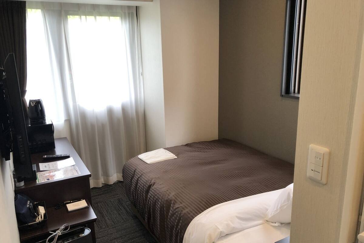 Ein Hotelzimmer mit einem Bett, einem Schreibtisch mit Computer und Kaffeemaschine, einem Fenster mit Vorhängen und einem an der Wand montierten Fernseher.