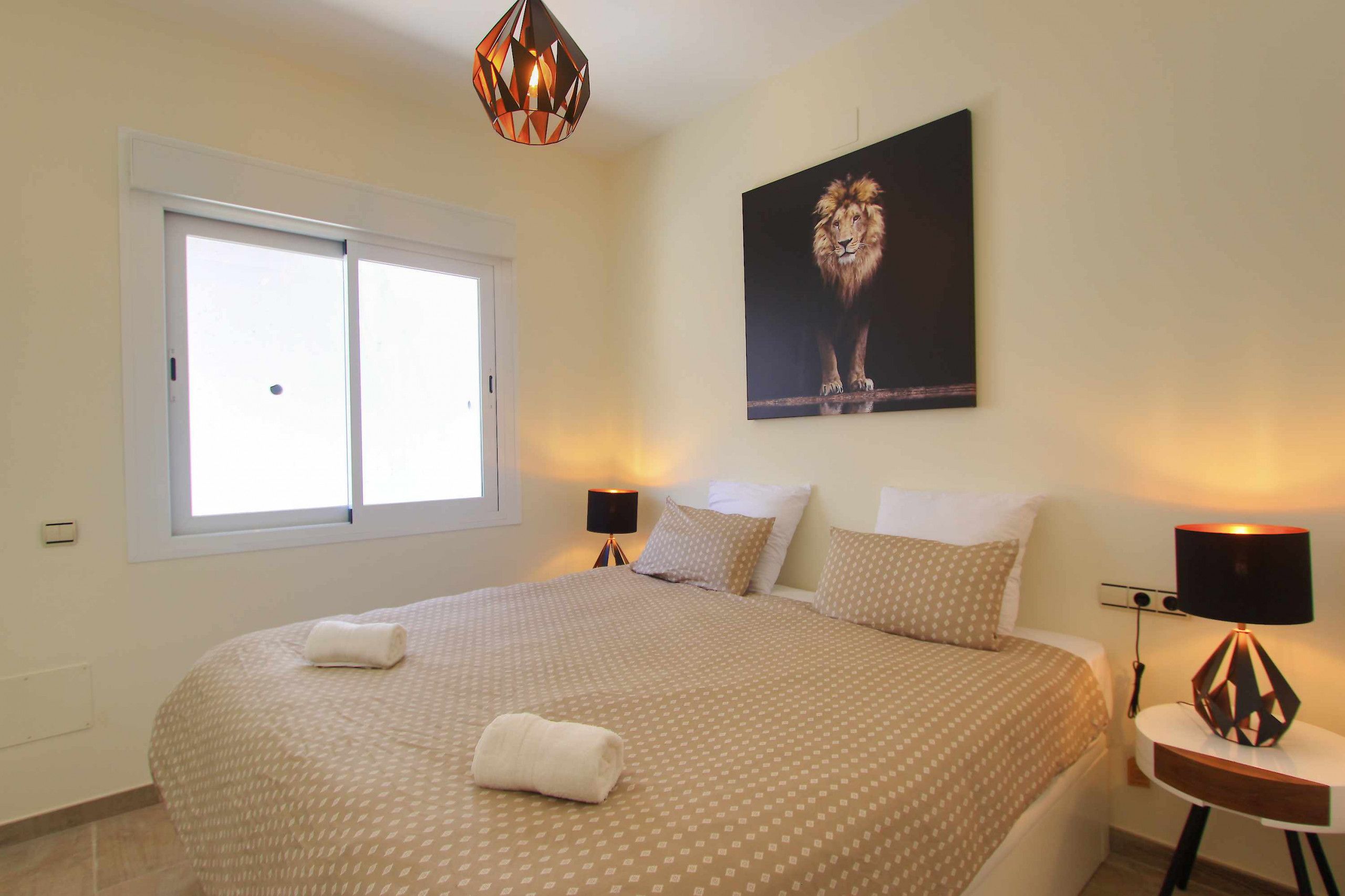 Quarto com uma cama grande, duas luminárias de cabeceira, uma janela e um quadro de leão na parede.