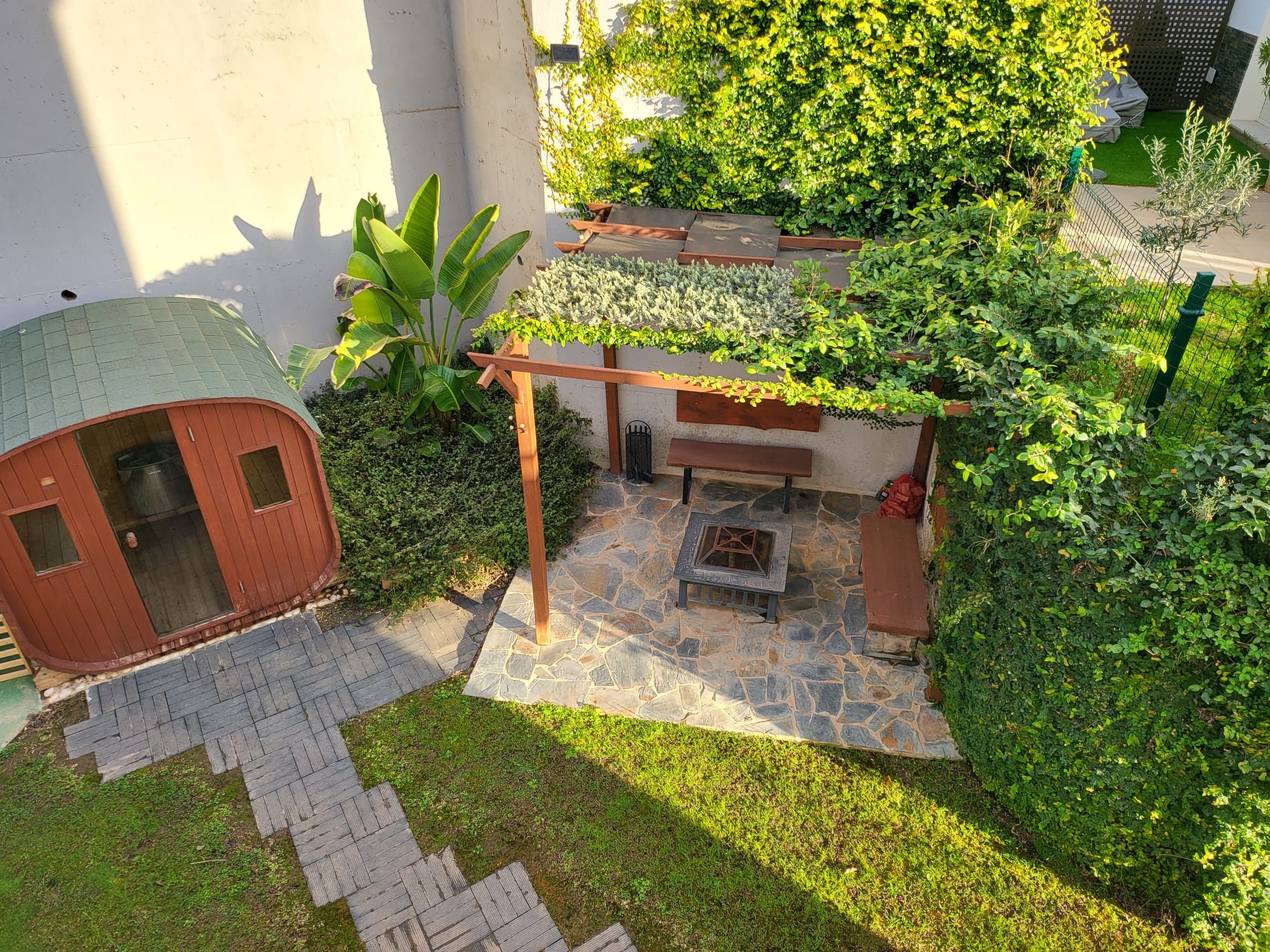 A patio area with a stone floor, wooden benches, a small fire pit, and a wooden pergola covered with climbing plants.