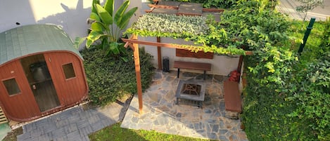 A patio area with a stone floor, wooden benches, a small fire pit, and a wooden pergola covered with climbing plants.