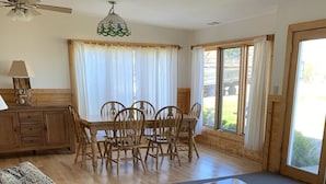 Ein Wohnzimmer mit einer Couch, einem Couchtisch, einem Esstisch mit Stühlen, einer Deckenleuchte und großen Fenstern mit Vorhängen.