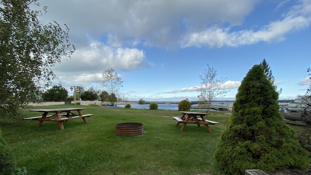 Ein Picknickplatz mit zwei Holztischen und -bänken, einer Feuerstelle aus Metall sowie Blick auf einen See mit Booten.