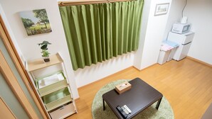A room with a wooden floor, a dark brown table, a green rug, a green curtain, a small plant on a shelf, a microwave, and a refrigerator. - Mell theater (Osaka)