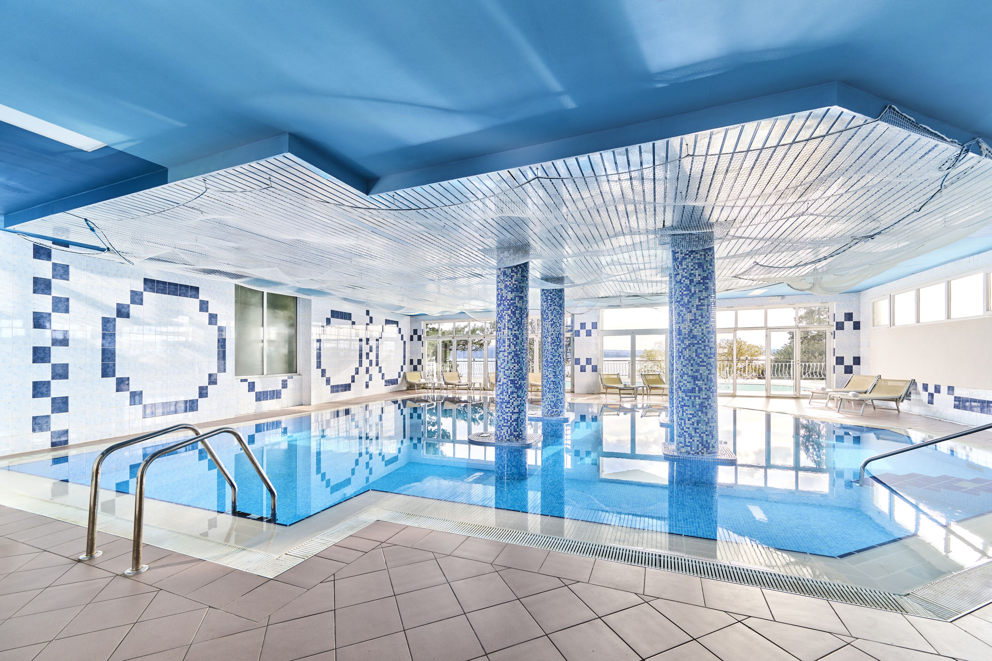 An indoor swimming pool with tiled walls and ceiling, clear glass barriers, and lounge chairs by the pool.