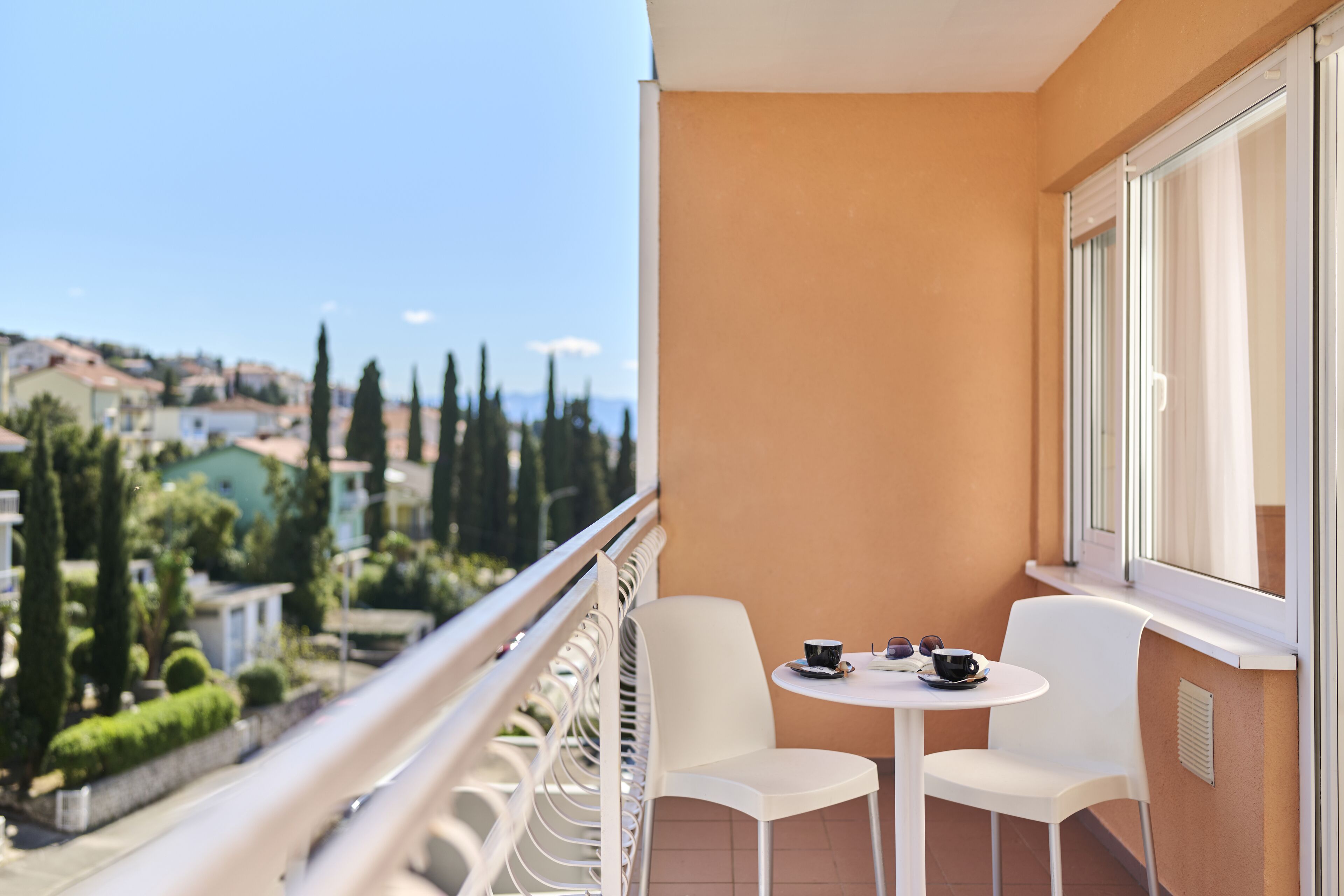 Un balcón con mesa y sillas blancas, con vistas a una zona residencial con casas y árboles.