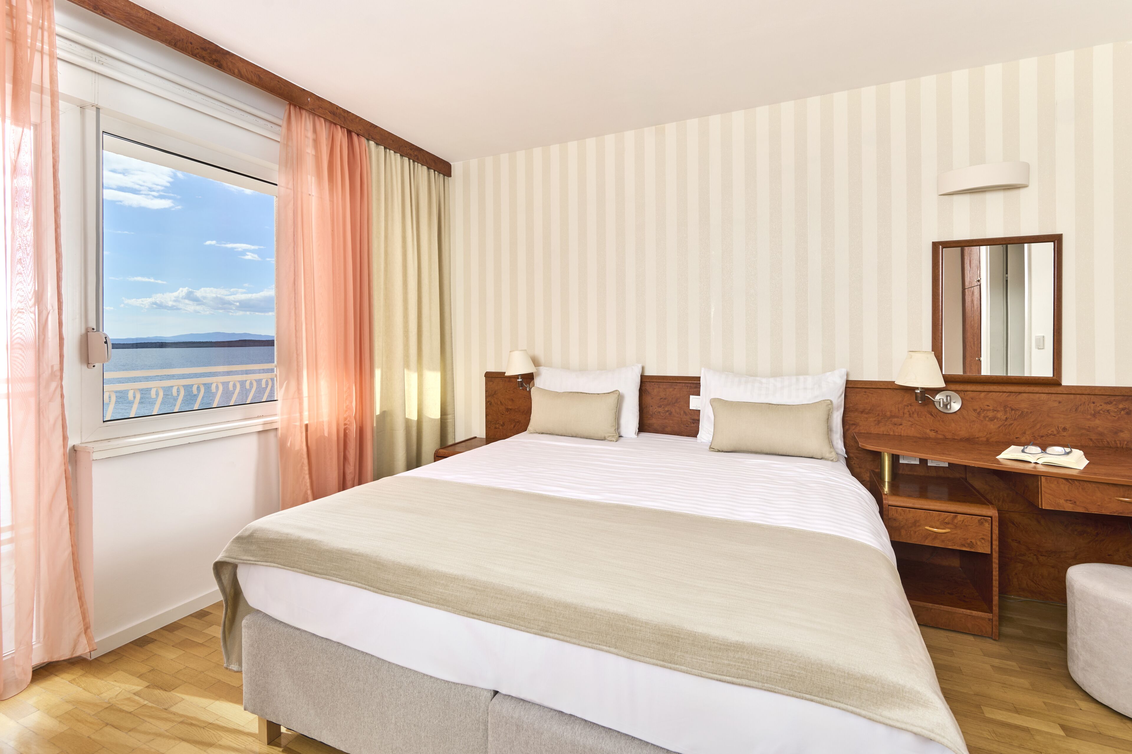 Une chambre d’hôtel avec un grand lit, un bureau avec un miroir et une vue sur la mer par la fenêtre.