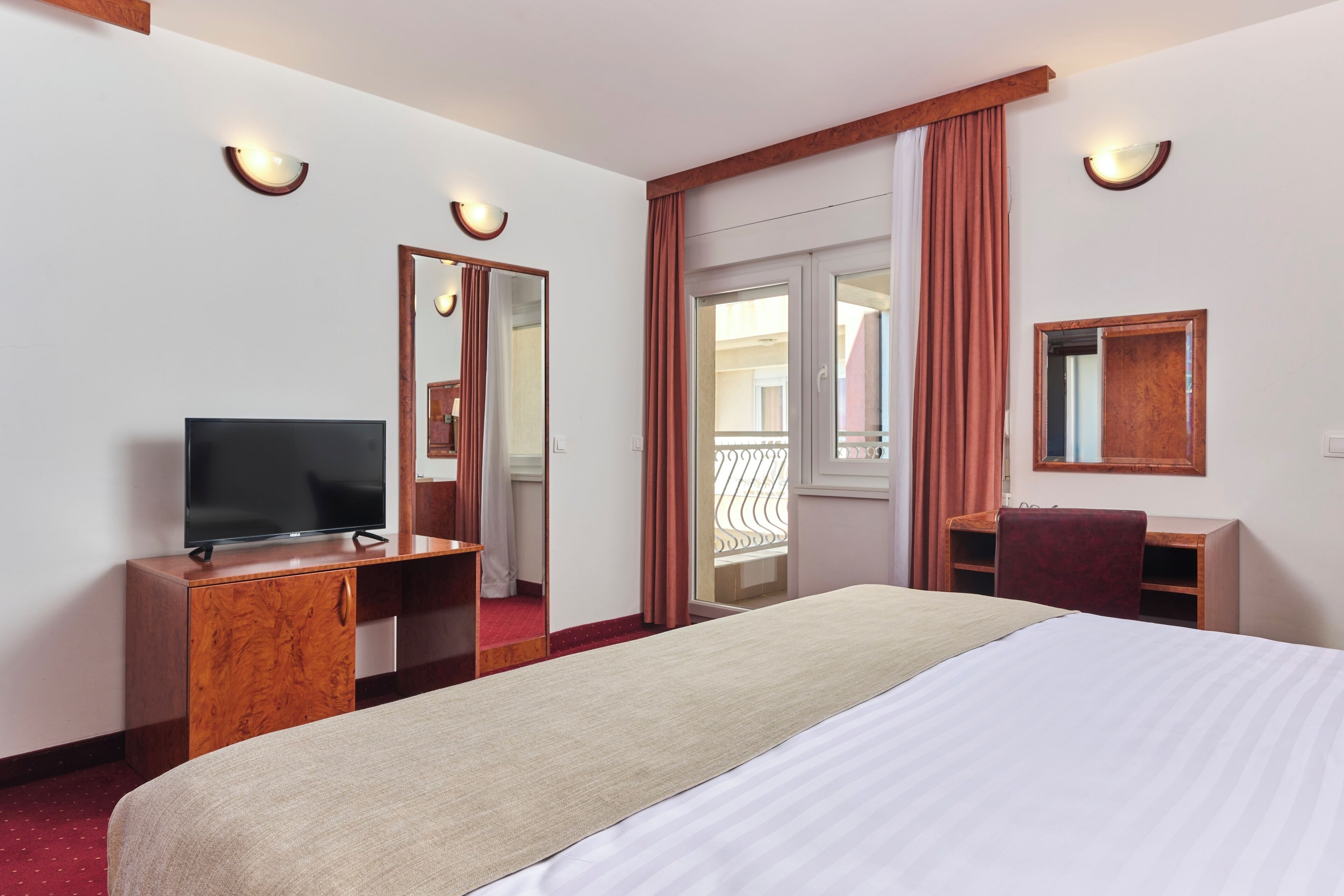A hotel room with a bed, a desk, a television, a mirror, and a sliding door to a balcony.