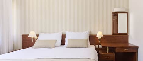 A hotel room with a large bed, two bedside tables with lamps, a wooden desk with a mirror, and a small stool.