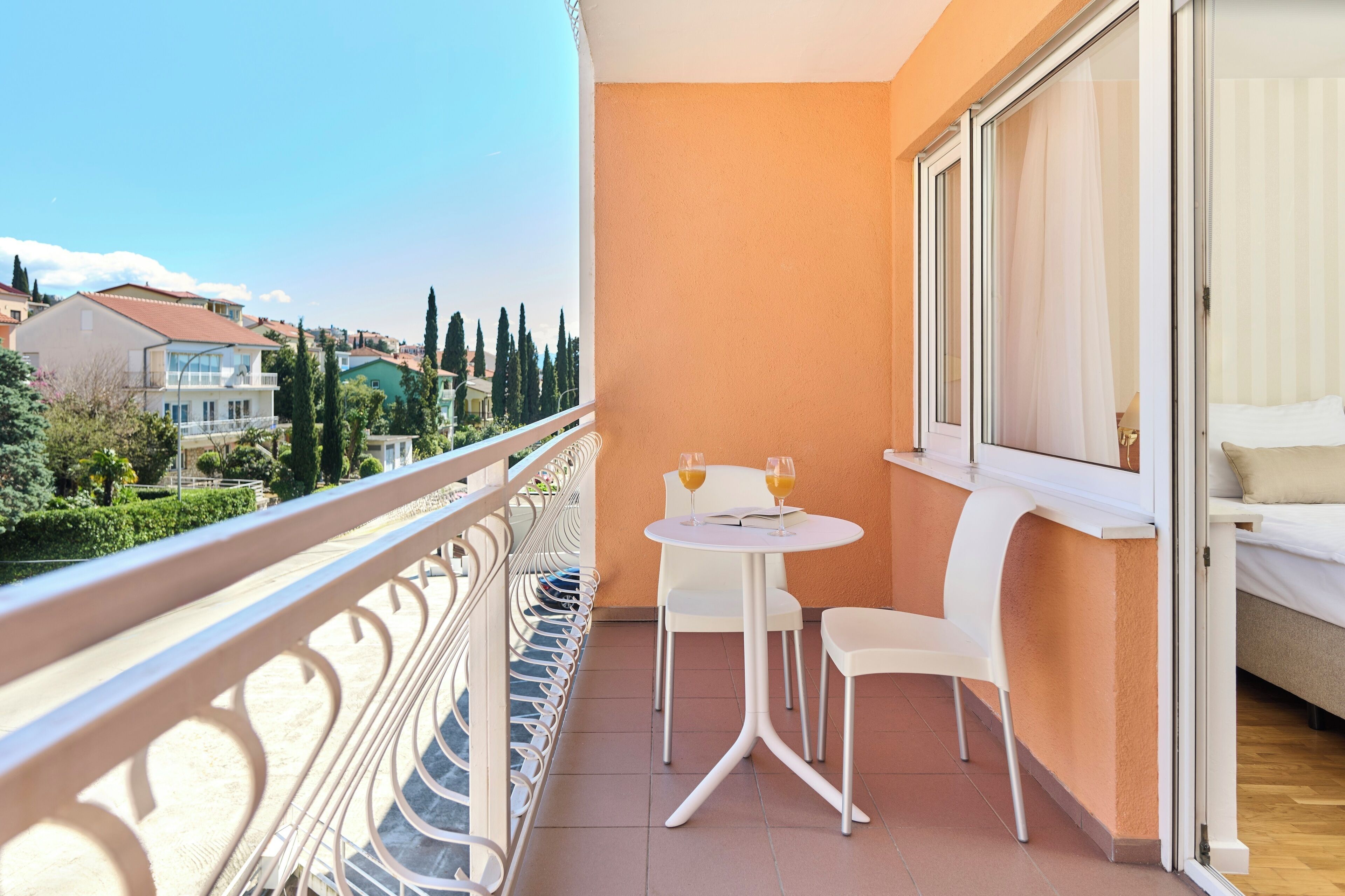 A balcony with a white table and chairs, a bed with a white headboard, and a view of the cityscape.