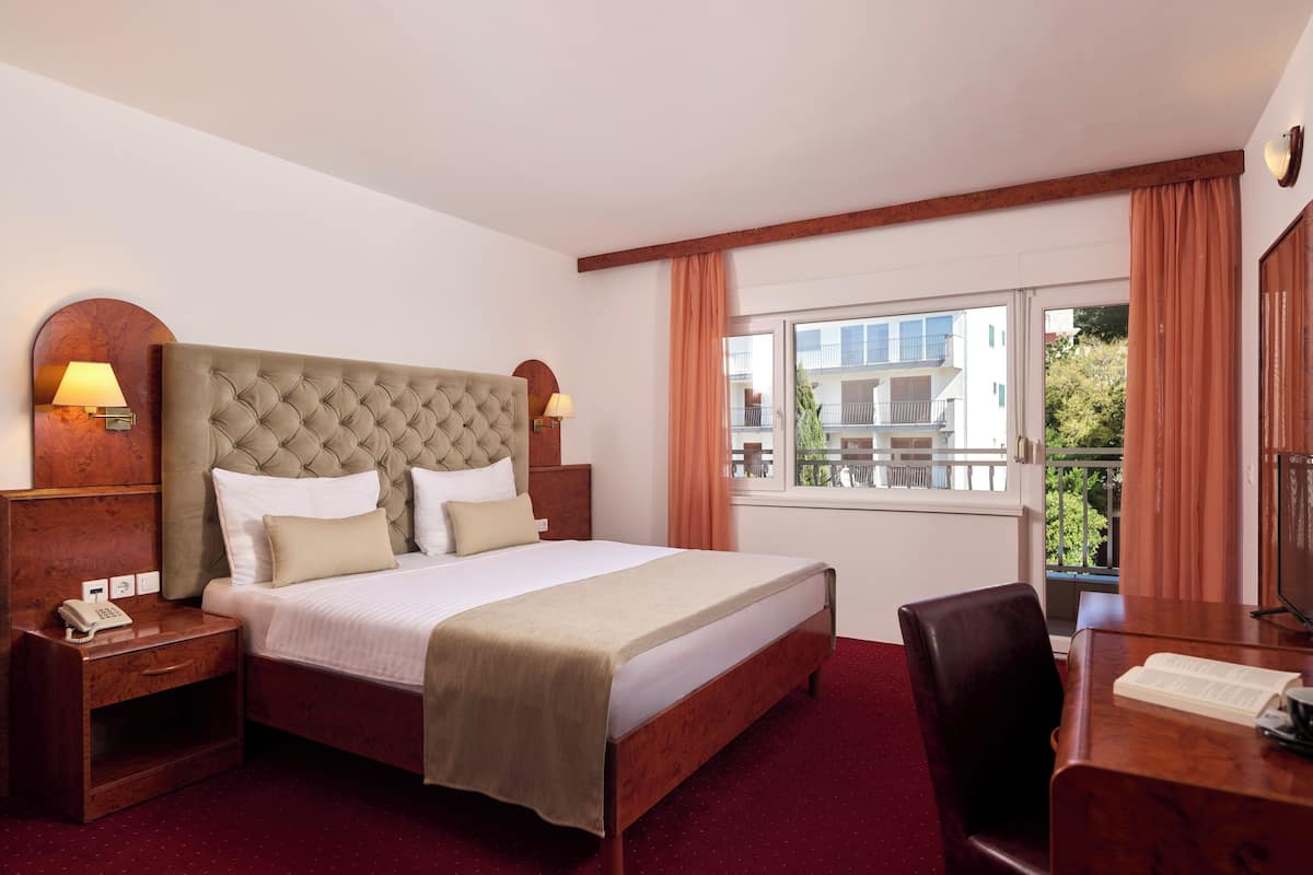 Une chambre d’hôtel avec un grand lit, une tête de lit en bois, deux lampes de chevet, une table de chevet en bois avec un téléphone, un bureau en bois avec une chaise et une fenêtre avec des rideaux.