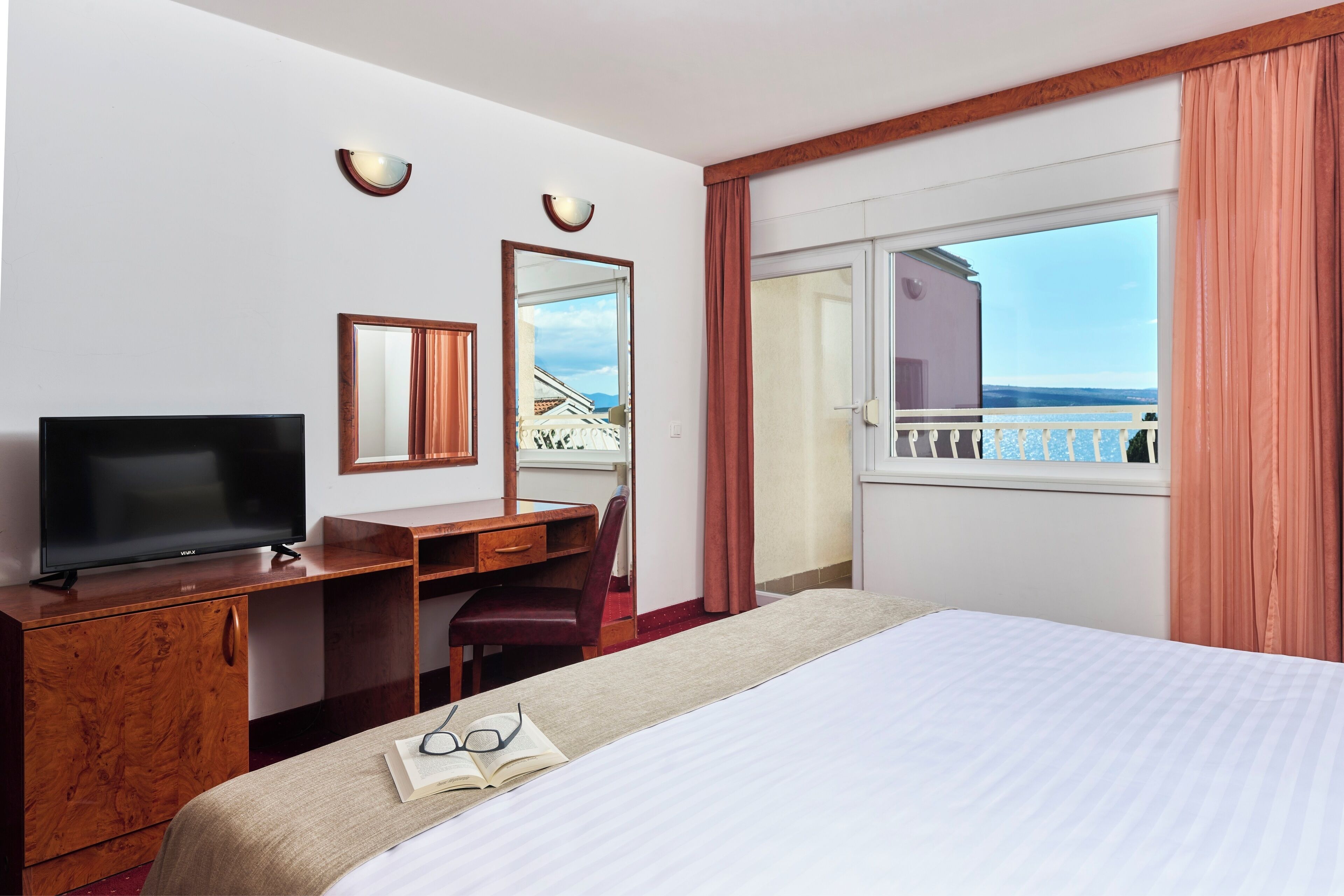 Habitación de hotel con cama, escritorio con televisor, silla, espejo y ventana con vista al balcón.