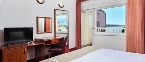 Habitación de hotel con cama, escritorio con televisor, silla, espejo y ventana con vista al balcón.