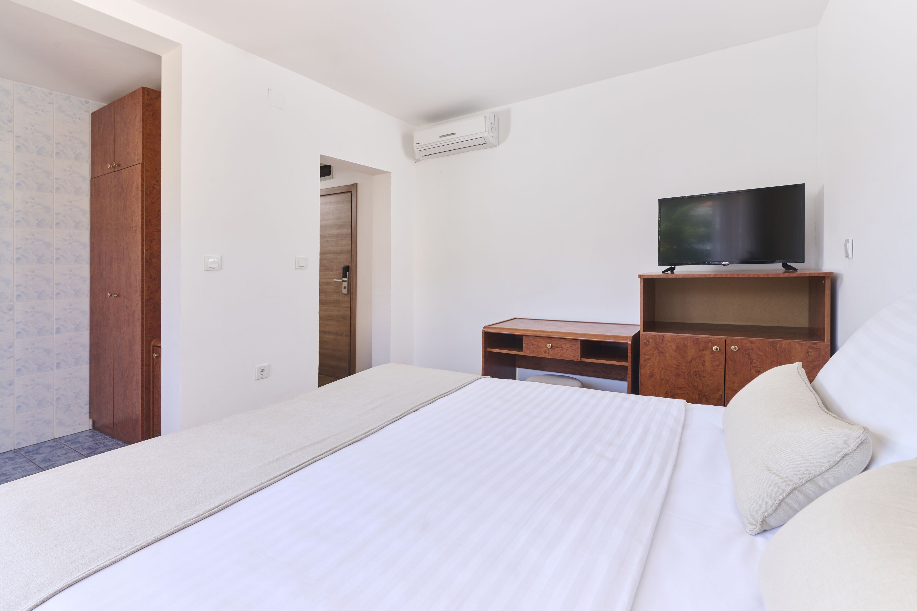 Ein Hotelzimmer mit einem Bett, einem Fernseher auf einem Holzständer, einem Kleiderschrank und einer Klimaanlage.