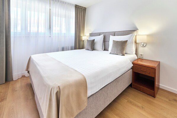 Una cama bien hecha con colcha y almohadas color beige, una mesita de noche de madera con una lámpara y cortinas traslúcidas en una ventana.