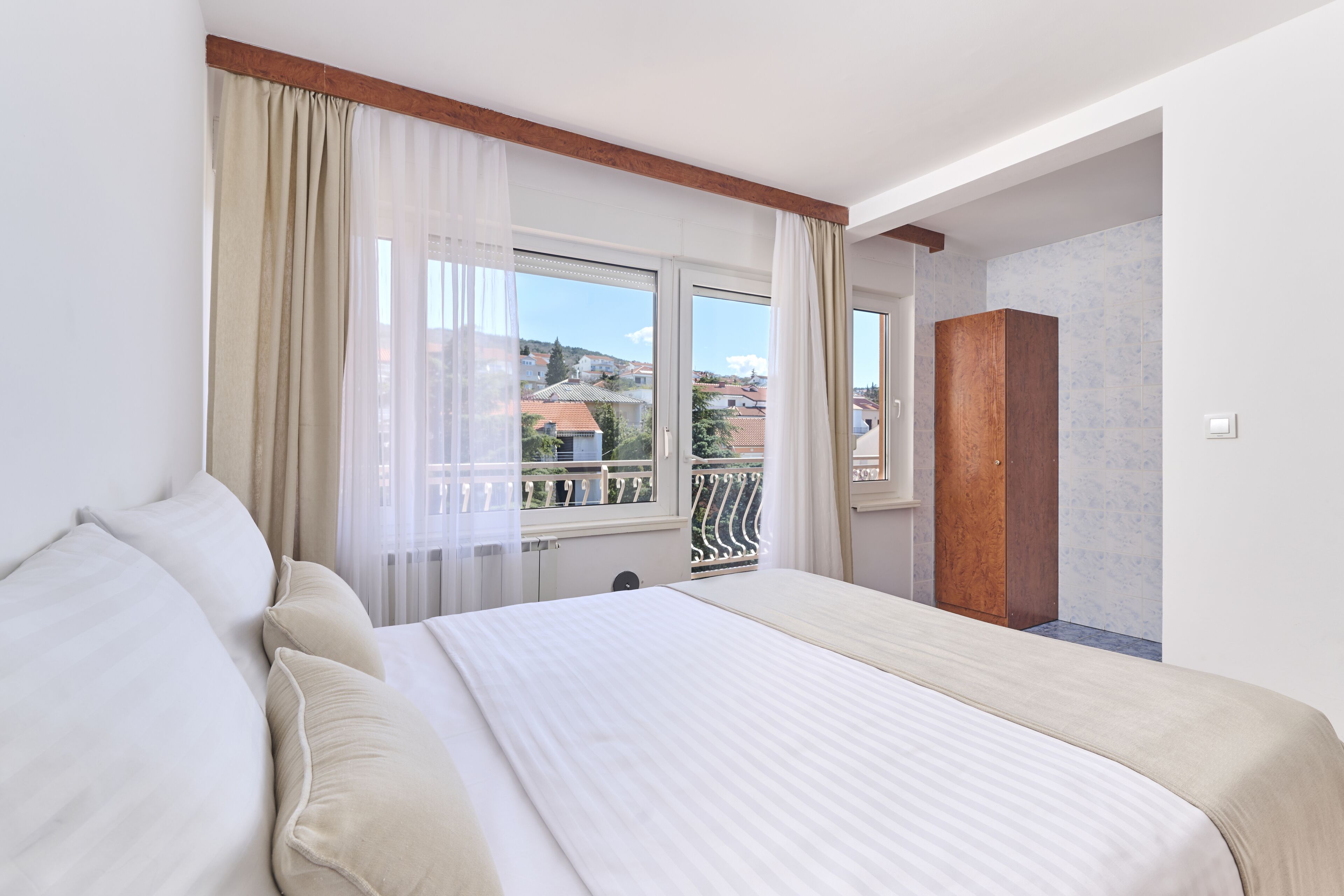 Une chambre d’hôtel avec un grand lit, une armoire en bois, un balcon donnant sur les immeubles et des rideaux légers à la fenêtre.
