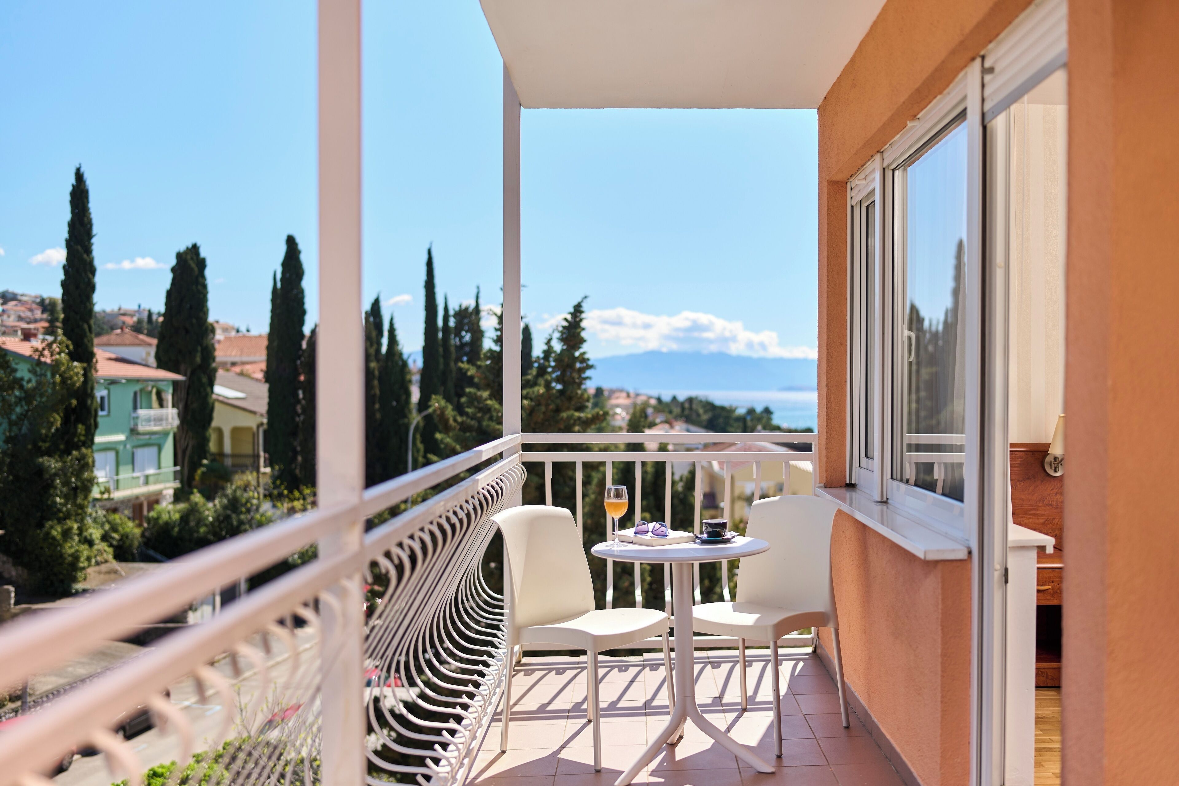 A balcony with a white railing, two white chairs, and a small table with a glass of wine.