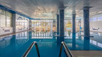 An indoor swimming pool with clear blue water, tiled walls, and columns with mosaic patterns.
