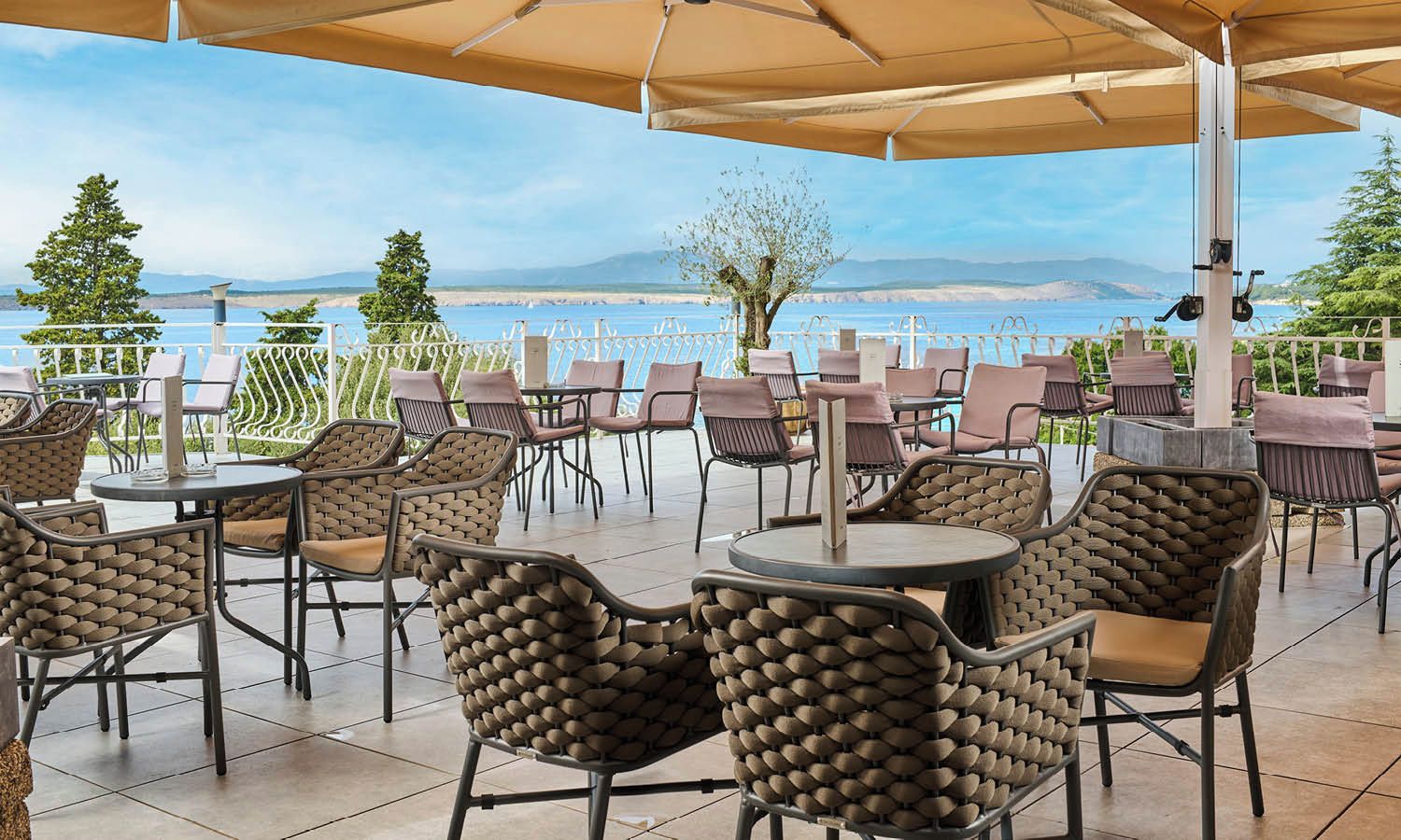 An outdoor seating area with round tables and wicker chairs, overlooking a body of water and distant mountains.