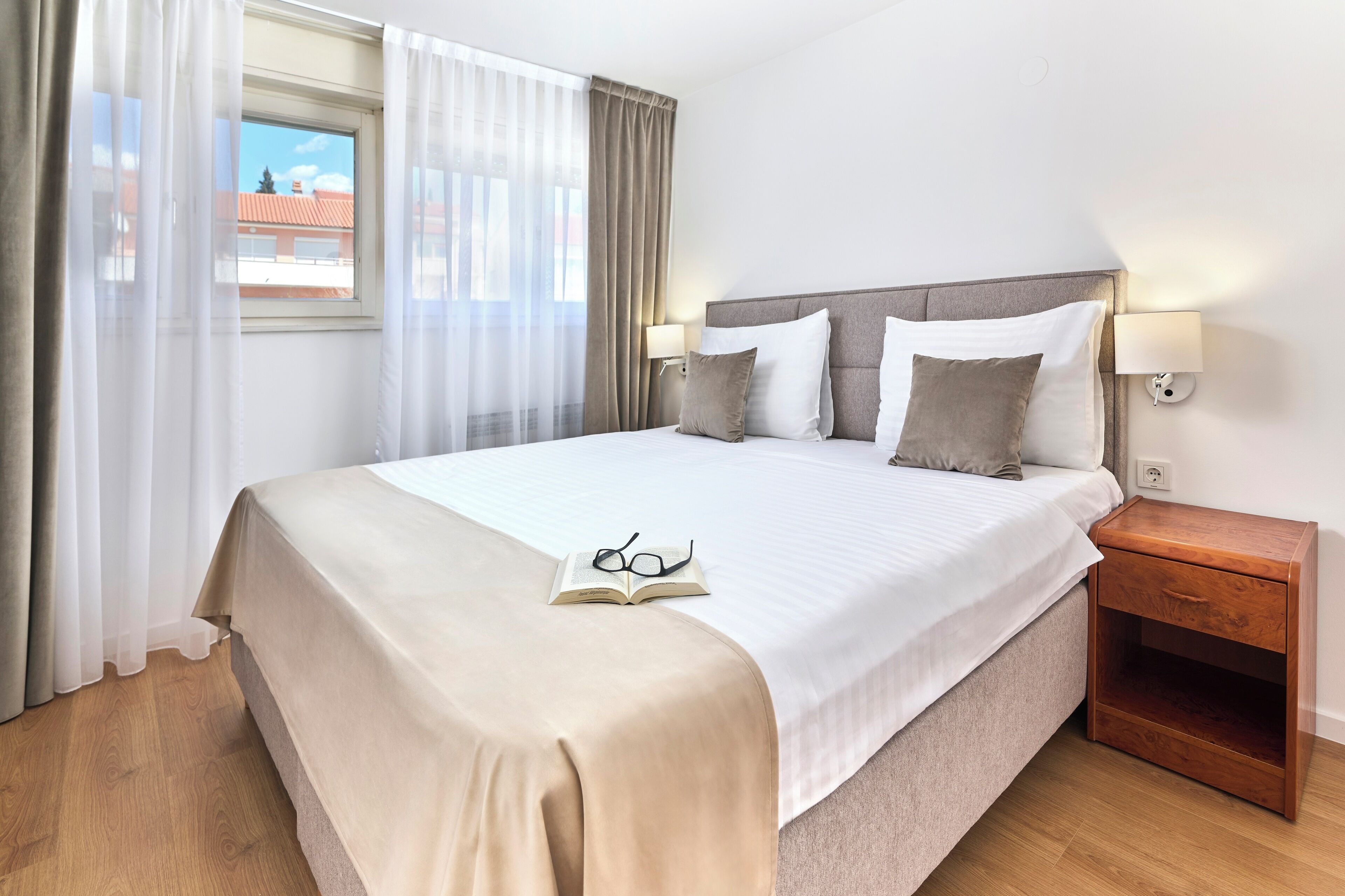 A neatly made bed with a beige bedspread, two pillows, and a pair of glasses on the bed. There is a wooden nightstand beside the bed with a lamp on it. The room has a large window with sheer curtains and a view of buildings outside.
