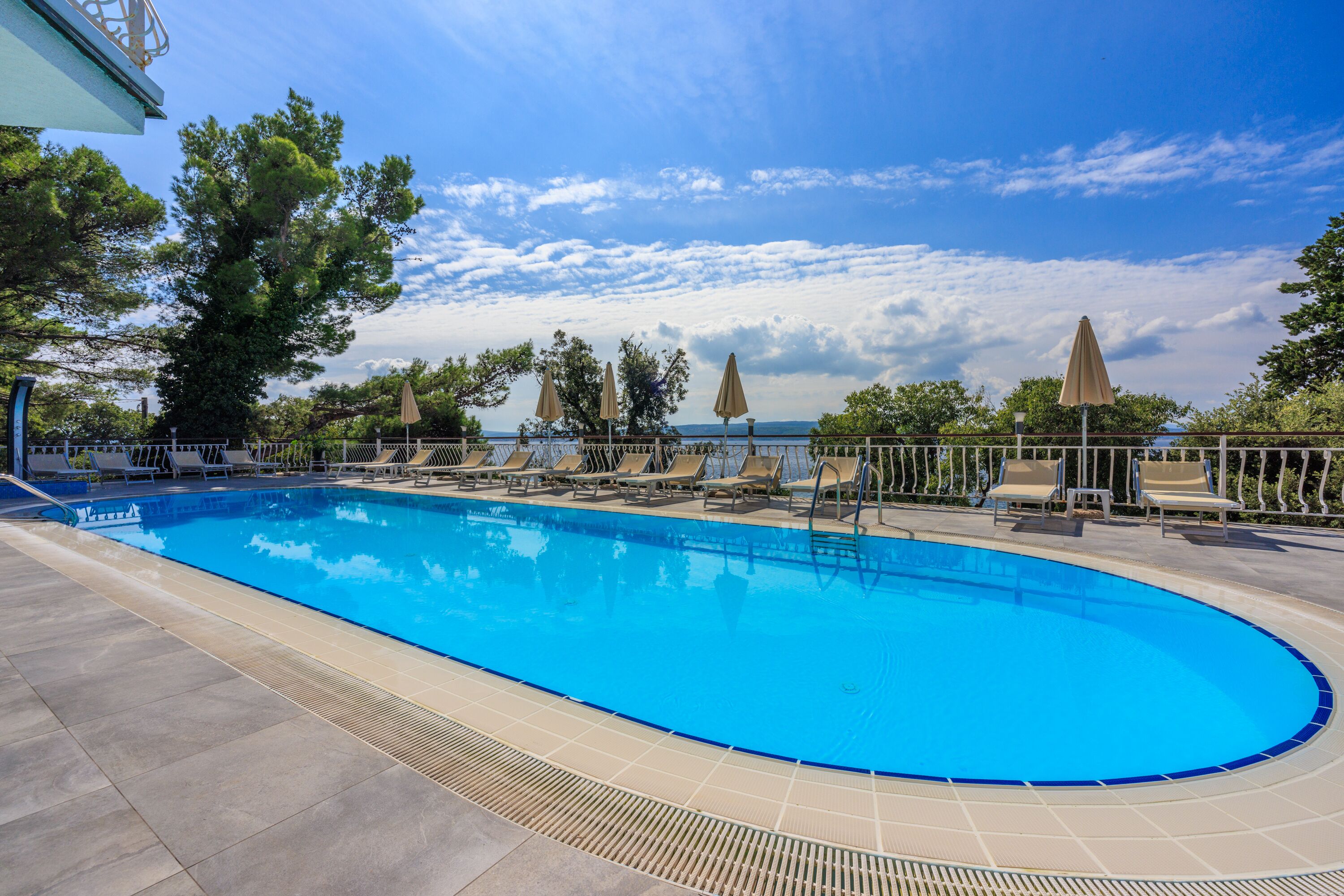 Ein Schwimmbecken mit Liegestühlen und Sonnenschirmen, umgeben von Bäumen und blauem Himmel.