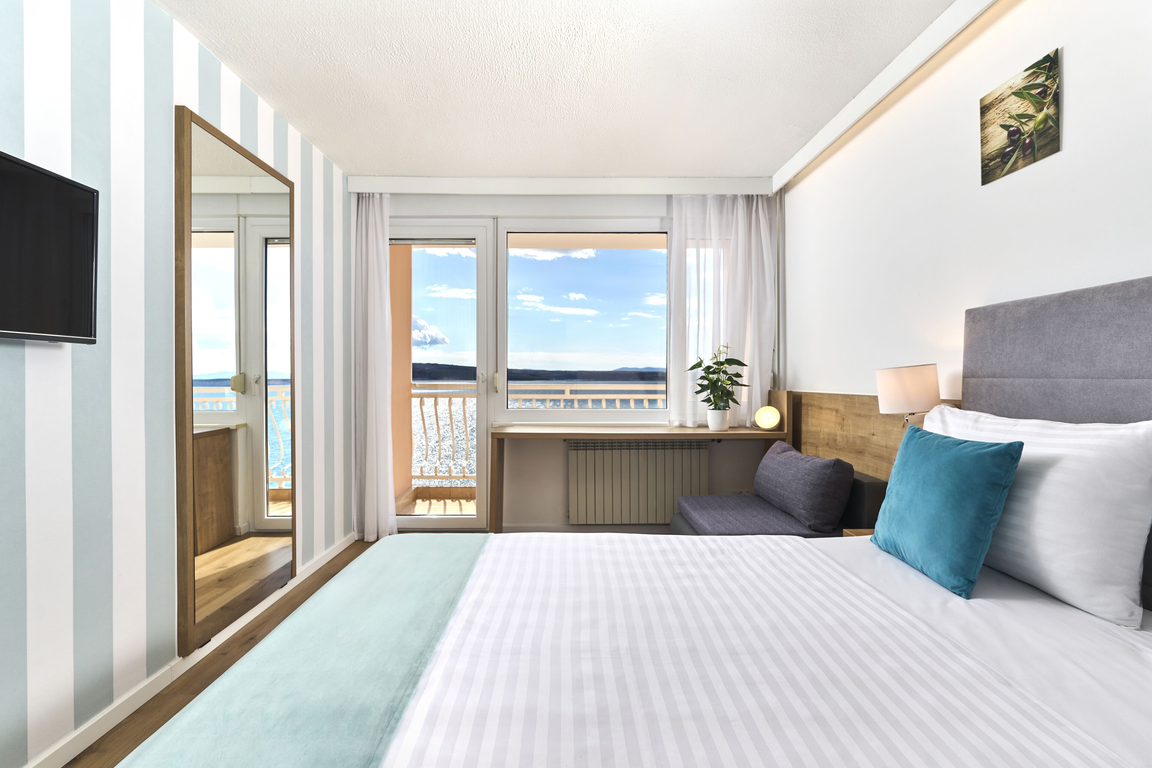 A hotel room with a large bed, a TV, a balcony with a view, and a plant on the bedside table.