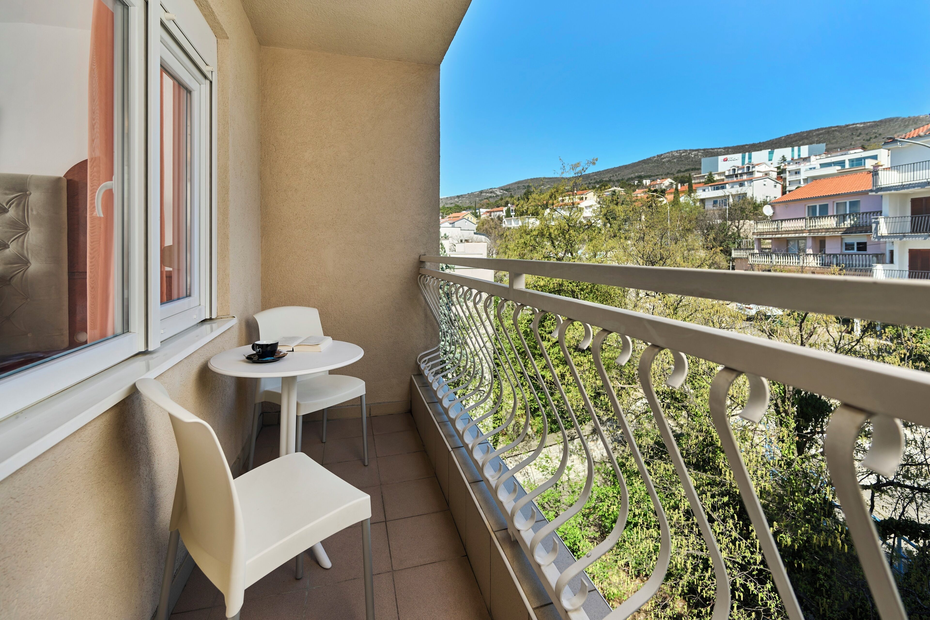 A balcony with a white railing, a small round table, two white chairs, and a window with curtains.
