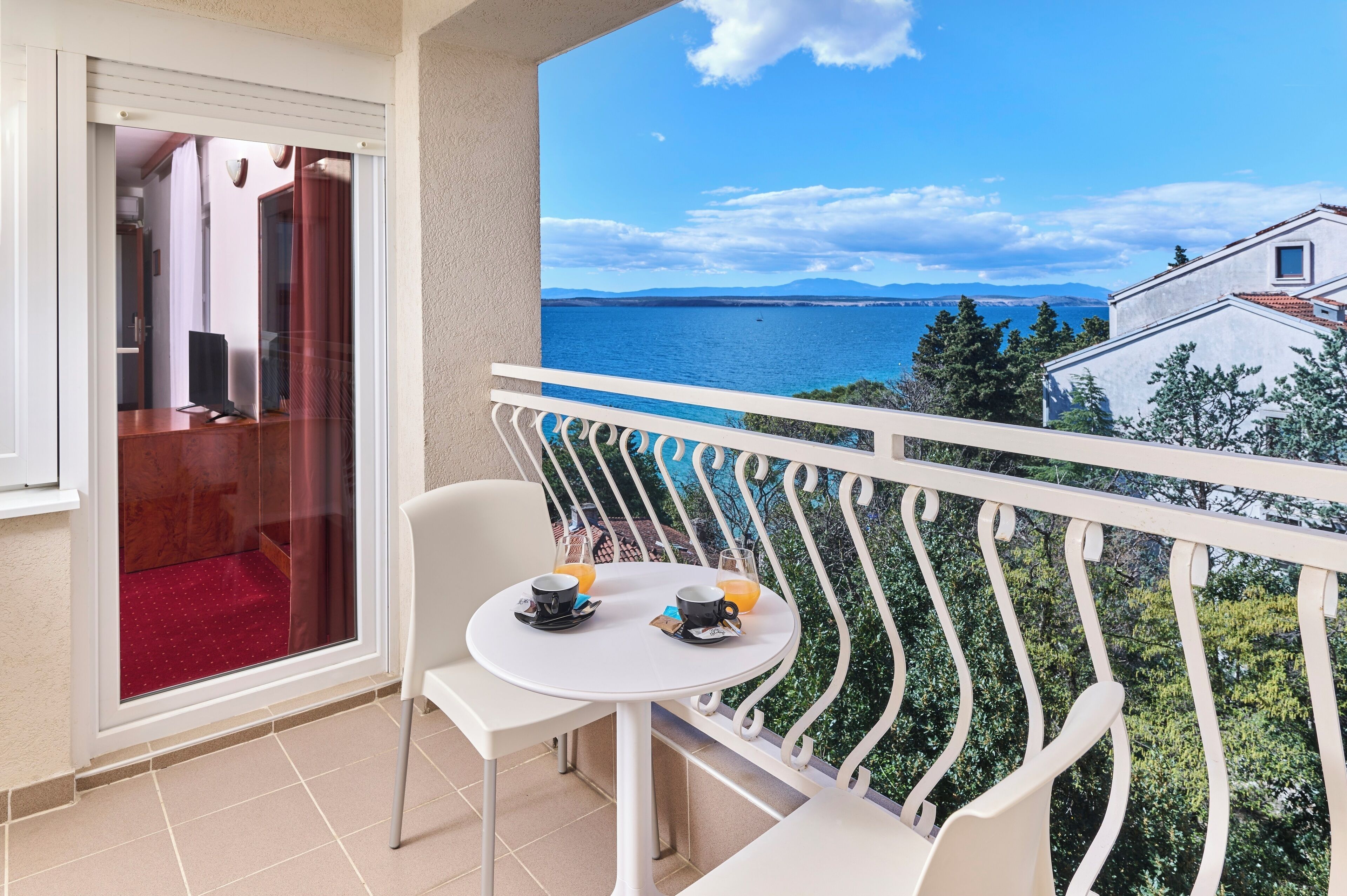 Un balcon avec une table et des chaises blanches, une porte vitrée et une vue sur la mer et les montagnes lointaines.