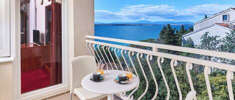 Un balcon avec une table et des chaises blanches, une porte vitrée et une vue sur la mer et les montagnes lointaines.